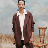 Male model with the 'Easy-Going Jacket' in brown linen with a beige shirt with large collars, and matching brown linen pants, shot on a light-blue textured background, waist-high beige grass, and a bamboo chair.