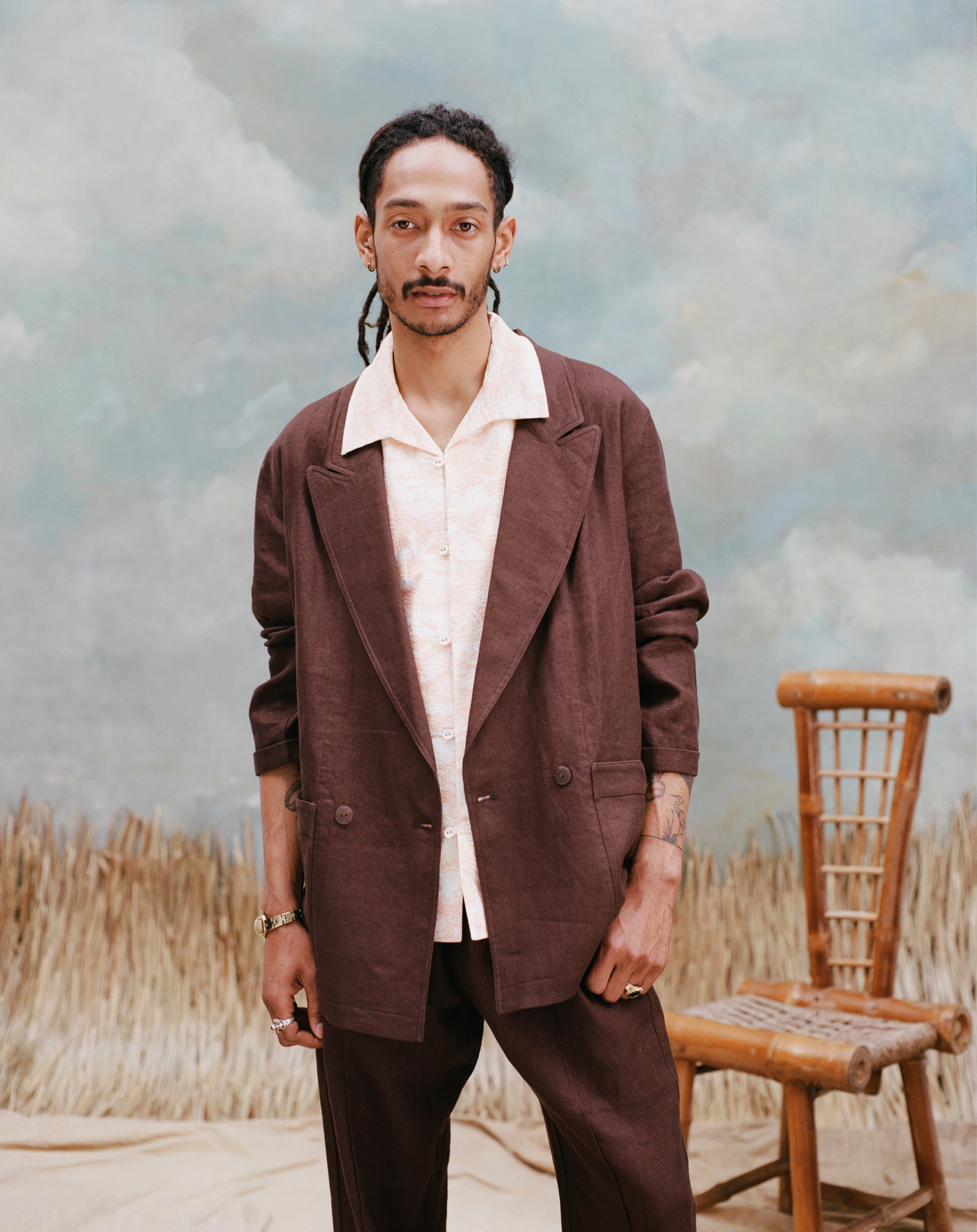 Male model with the 'Easy-Going Jacket' in brown linen with a beige shirt with large collars, and matching brown linen pants, shot on a light-blue textured background, waist-high beige grass, and a bamboo chair.