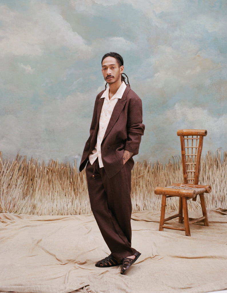Male model with dreadlocks wearing the 'Easy Going Jacket' in brown linen with matching brown linen pants, brown sandals, and a white button down shirt with large collars, shot on a light blue textured background, waist high beige grass, and a bamboo chair.