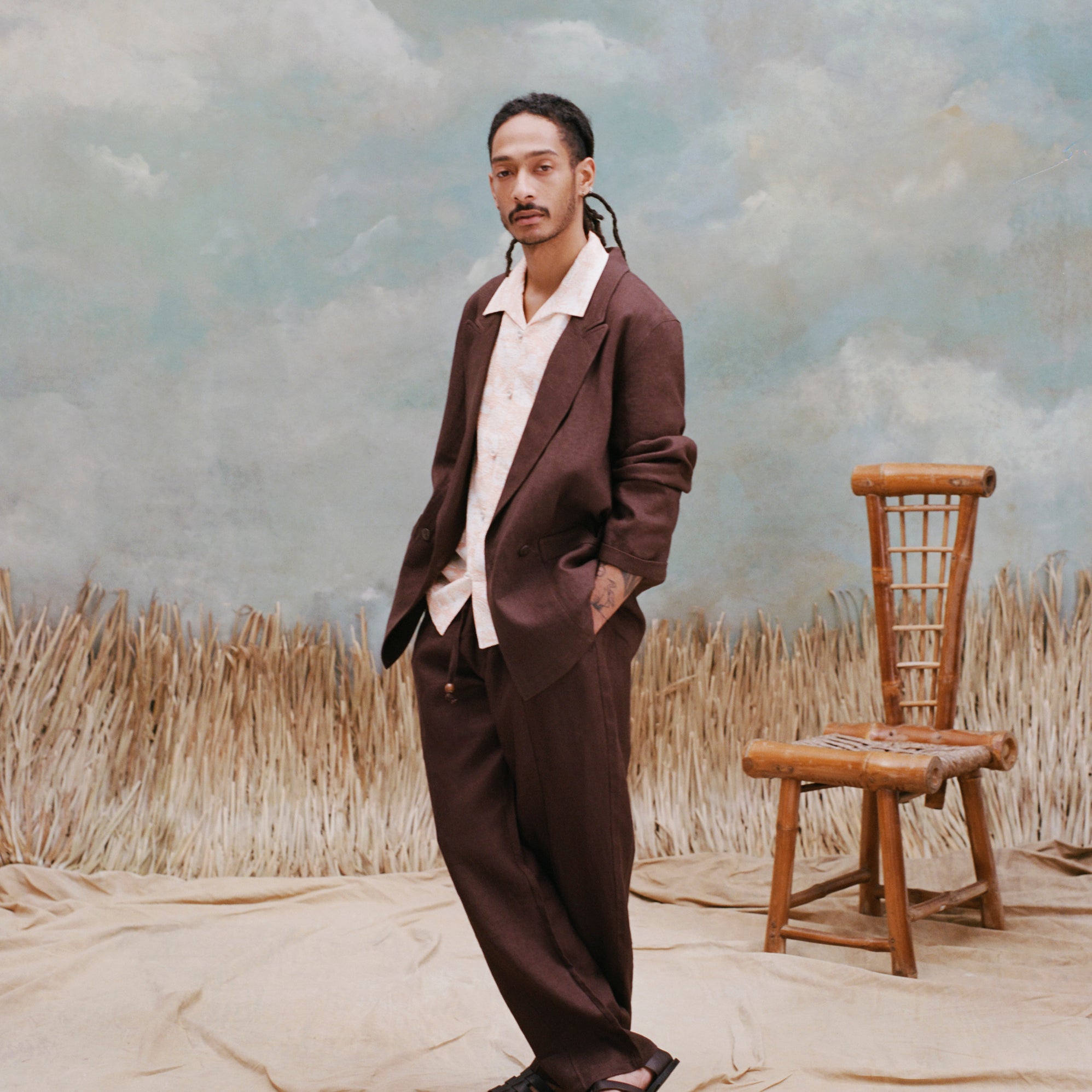 Male model with dreadlocks wearing the 'Easy-Going Pants' in brown linen with abrown blazer, white button-down shirt, and brown sandals, shot on a textured light blue background with a bamboo chair, and beige waist-high grass.