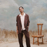 Male model with dreadlocks wearing the 'Easy-Going Jacket' in brown linen unbuttoned with a white buttoned down shirt with large collars, matching brown linen pants, and brown sandals, shot on a background with light blue texturing, waist high beige grass, and a bamboo chair.