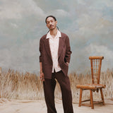 Male model with dreadlocks wearing the 'Easy-Going Jacket' in brown linen unbuttoned with a white buttoned down shirt with large collars, matching brown linen pants, and brown sandals, shot on a background with light blue texturing, waist high beige grass, and a bamboo chair.
