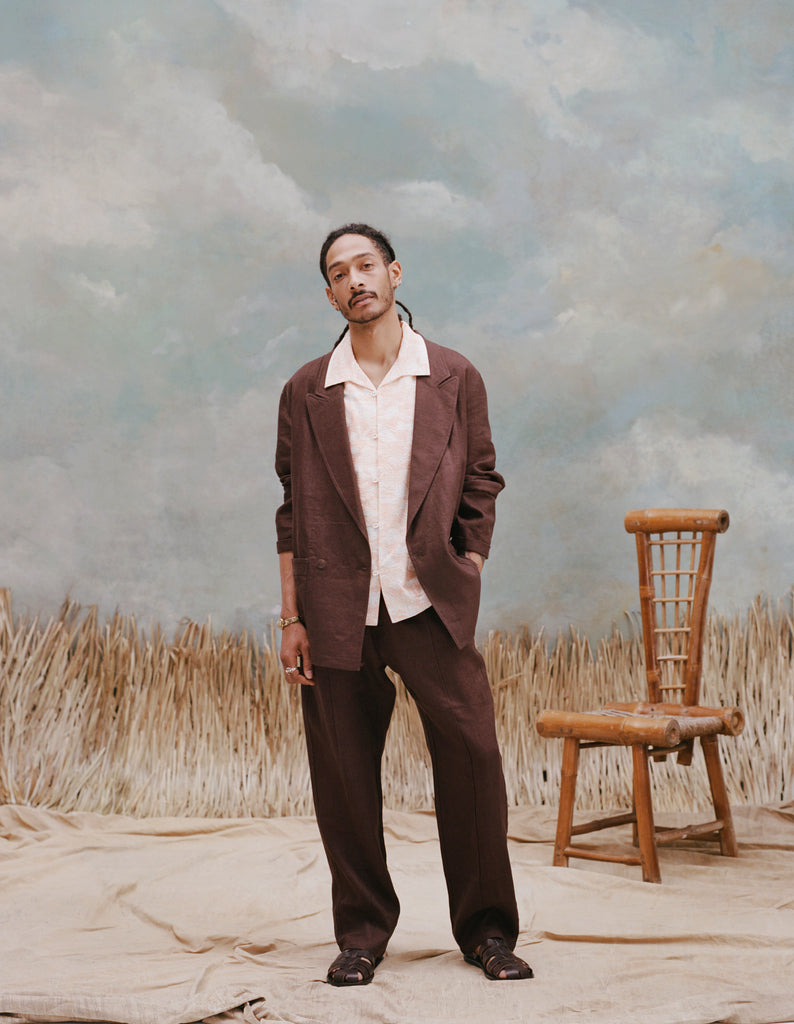 Male model with dreadlocks wearing the 'Easy-Going Jacket' in brown linen unbuttoned with a white buttoned down shirt with large collars, matching brown linen pants, and brown sandals, shot on a background with light blue texturing, waist high beige grass, and a bamboo chair.