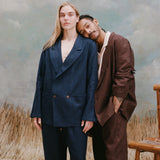 Female model with long blonde hair wearing the 'easy-going jacket' in navy linen alongside matching navy linen pants, and a male model with dreadlocks resting his head on the female model shoulder's while wearing an easy-going jacket in brown linen and matching brown linen pants, shot on a light blue textured background with beige grass.