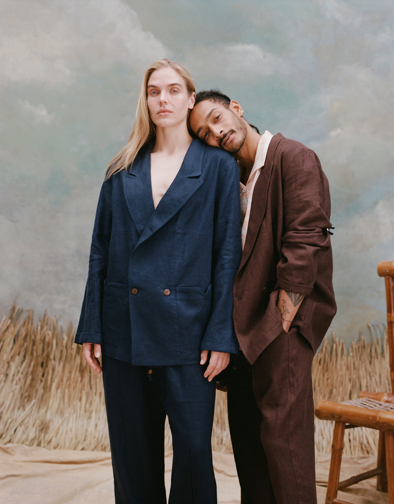 Female model with long blonde hair wearing the 'easy-going jacket' in navy linen alongside matching navy linen pants, and a male model with dreadlocks resting his head on the female model shoulder's while wearing an easy-going jacket in brown linen and matching brown linen pants, shot on a light blue textured background with beige grass.