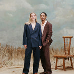 female model with long blonde hair wearing the 'easy-going' jacket in navy linen buttoned with matching navy linen pants and light brown sandals with a male model with dreadlocks standing slight beside and behind the female model while wearing the easy-going jacket in brown linen with a white cabana shirt underneath and matching brown linen pants with brown sandals, shot on a light blue textured background with beige grass.