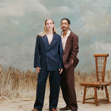 female model with long blonde hair wearing the 'easy-going' jacket in navy linen buttoned with matching navy linen pants and light brown sandals with a male model with dreadlocks standing slight beside and behind the female model while wearing the easy-going jacket in brown linen with a white cabana shirt underneath and matching brown linen pants with brown sandals, shot on a light blue textured background with beige grass.