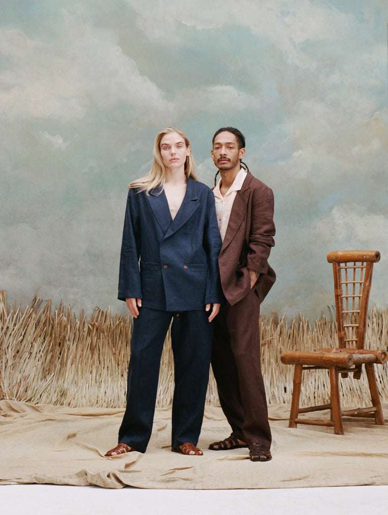 female model with long blonde hair wearing the 'easy-going' jacket in navy linen buttoned with matching navy linen pants and light brown sandals with a male model with dreadlocks standing slight beside and behind the female model while wearing the easy-going jacket in brown linen with a white cabana shirt underneath and matching brown linen pants with brown sandals, shot on a light blue textured background with beige grass.