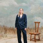 Female model with long-blonde hair wearing the "easy-going jacket' in navy linen with matching navy linen pants and brown sandals, shot on a light blue textured background with beige grass and a bamboo chair.