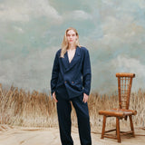 Female model with long-blonde hair wearing the "easy-going jacket' in navy linen with matching navy linen pants and brown sandals, shot on a light blue textured background with beige grass and a bamboo chair.