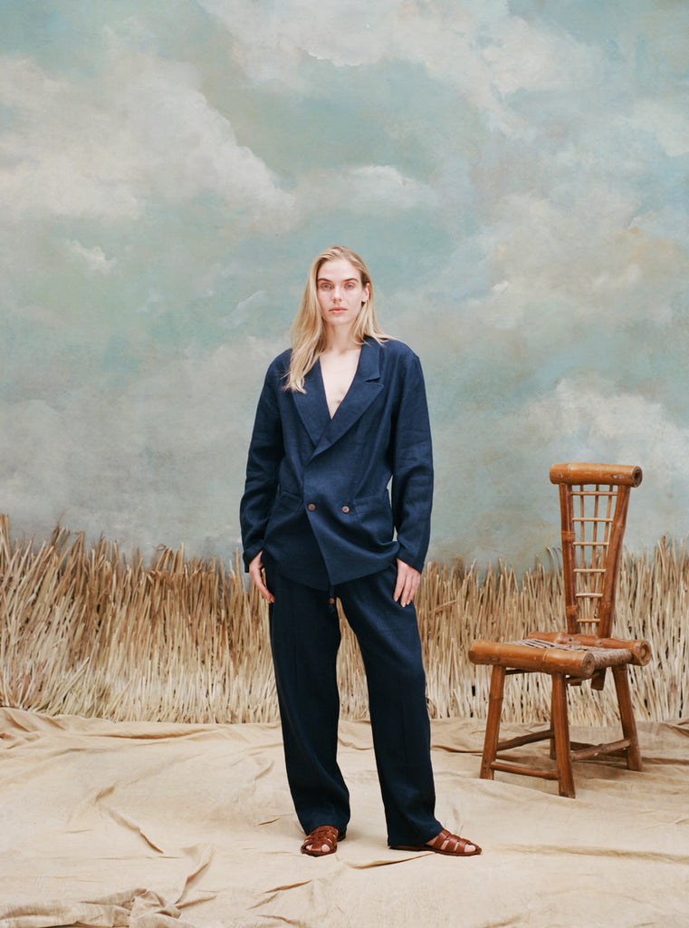 Female model with long-blonde hair wearing the "easy-going jacket' in navy linen with matching navy linen pants and brown sandals, shot on a light blue textured background with beige grass and a bamboo chair.