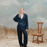 Female model with long-blonde hair wearing the 'Easy-Going Jacket' in navy linen with navy linen matching pants and brown sandals, shot on a light blue textured background with beige waist-high grass and a bamboo chair.