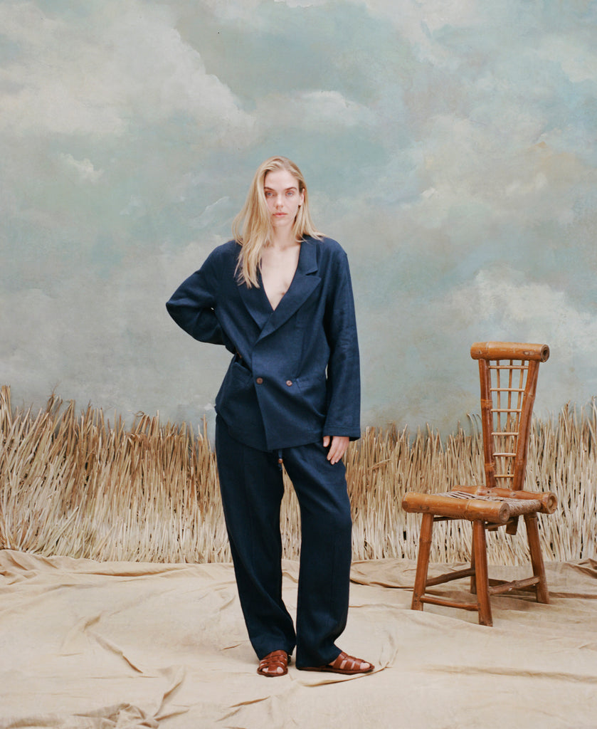Female model with long-blonde hair wearing the 'Easy-Going Jacket' in navy linen with navy linen matching pants and brown sandals, shot on a light blue textured background with beige waist-high grass and a bamboo chair.