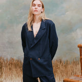 Female model with long blonde hair wearing the 'Easy-Going Jacket' in navy linen with navy linen pants, shot on a light blue textured background with waist-high beige grass and a bamboo chair.