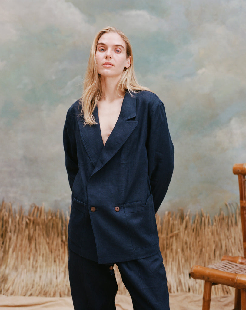 Female model with long blonde hair wearing the 'Easy-Going Jacket' in navy linen with navy linen pants, shot on a light blue textured background with waist-high beige grass and a bamboo chair.