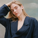 Female model with long blonde hair wearing the 'Easy-Going Jacket' in navy linen with one hand running through her hair, shot on a light blue textured background.