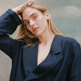 Female model with long blonde hair wearing the 'Easy-Going Jacket' in navy linen with one hand running through her hair, shot on a light blue textured background.