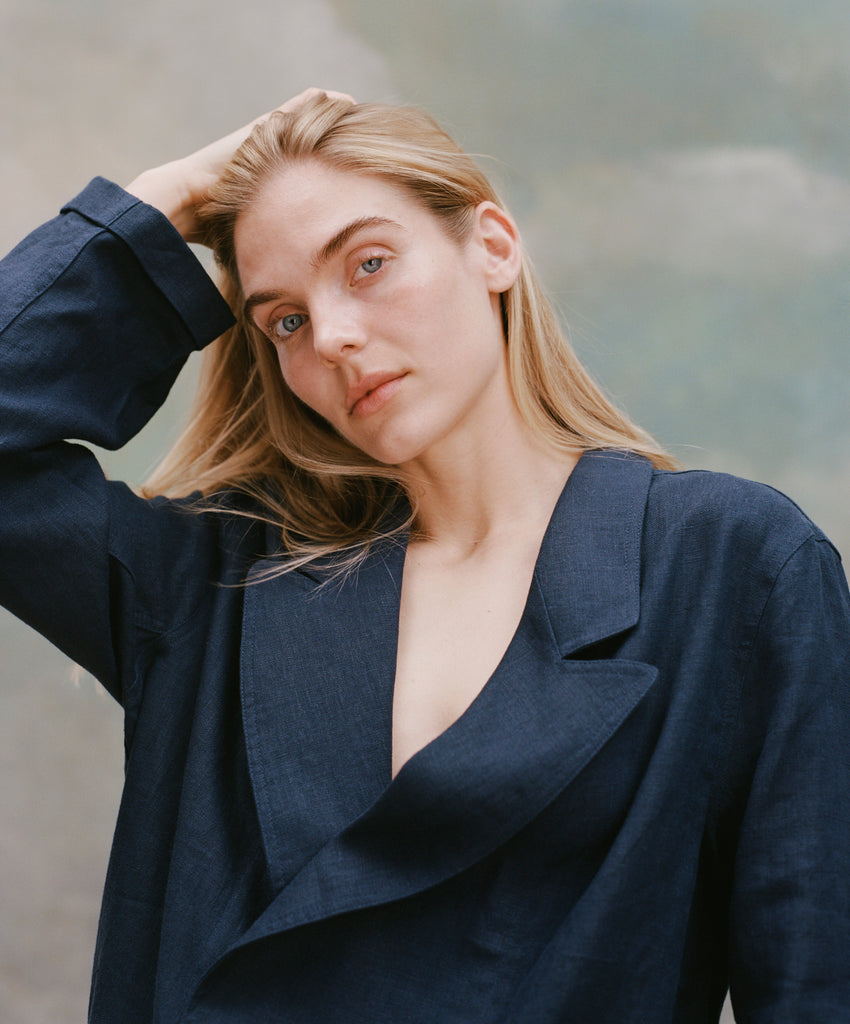 Female model with long blonde hair wearing the 'Easy-Going Jacket' in navy linen with one hand running through her hair, shot on a light blue textured background.