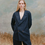 Female model with long-blonde hair wearing the 'Easy Going Jacket' in navy linen with matching navy linen pants, shot on a light blue textured background with waist high beige grass.