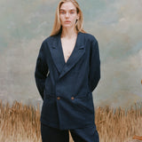 Female model with long-blonde hair wearing the 'Easy Going Jacket' in navy linen with matching navy linen pants, shot on a light blue textured background with waist high beige grass.