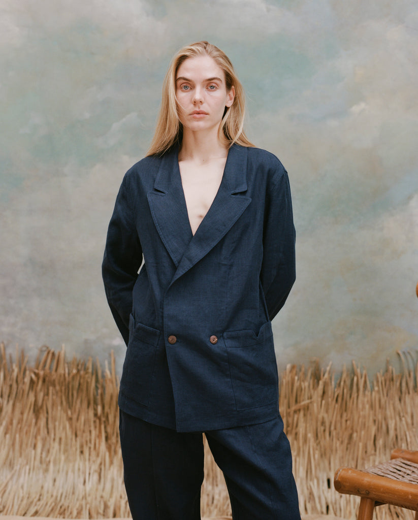 Female model with long-blonde hair wearing the 'Easy Going Jacket' in navy linen with matching navy linen pants, shot on a light blue textured background with waist high beige grass.