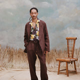 Male model with dreadlocks wearing the 'Easy-Going Jacket' in brown linen with a brown shirt featuring hibiscus and palm frond patterning, matching brown linen pants, shot on a light blue textured background with waist high grass and a bamboo chair.