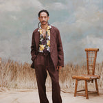 Young male model with one hand in their pocket wearing "Night Bloom" Shirt with one button undone and Easy-going Jacket and Easy-going pants in brown as well as black sandels with a light blue background that features wild grass and a wooden chair.