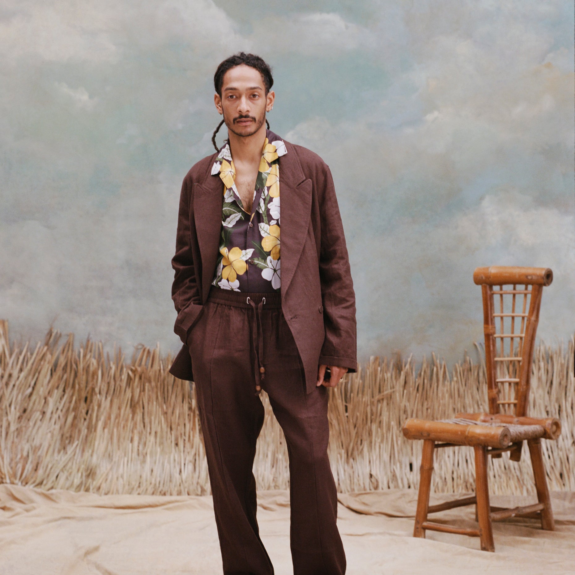 Male model with dreads wearing the 'Easy-Going Pants' in brown linen with a brown blazer and brown button down shirt with patterns of palm fronds and hibiscus, shot a on textured light blue background, beige waist-high grass, and a bamboo chair.