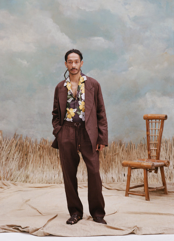 Male model with dreads wearing the 'Easy-Going Pants' in brown linen with a brown blazer and brown button down shirt with patterns of palm fronds and hibiscus, shot a on textured light blue background, beige waist-high grass, and a bamboo chair.