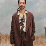 Male model with dreadlocks wearing the 'Easy-Going Jacket' in brown linen unbuttoned with a brown buttom shirt with a palm and hibiscus pattern, and matching brown linen pants with light-blue texturing, waist-high beige sand, and a bamboo chair.