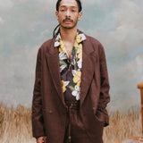 Male model with dreadlocks wearing the 'Easy-Going Jacket' in brown linen unbuttoned with a brown buttom shirt with a palm and hibiscus pattern, and matching brown linen pants with light-blue texturing, waist-high beige sand, and a bamboo chair.