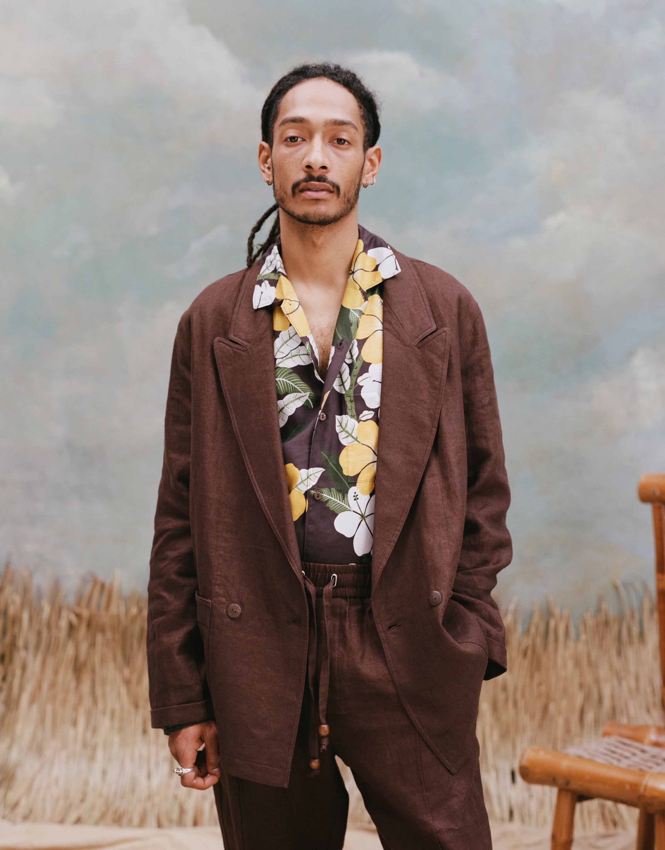 Male model with dreadlocks wearing the 'Easy-Going Jacket' in brown linen unbuttoned with a brown buttom shirt with a palm and hibiscus pattern, and matching brown linen pants with light-blue texturing, waist-high beige sand, and a bamboo chair.