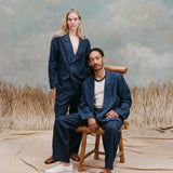 Female model with long-blonde hair next to a seated male model with dreadlocks as they both wear the 'Easy-Going Jacket' in Navy Linen alongside matching navy pants, with the male wearing a white shirt underneath and white sneakers while the female model is wearing nothing underneath the jacket, all shot on a textured light-blue background with waist-high beige grass. 