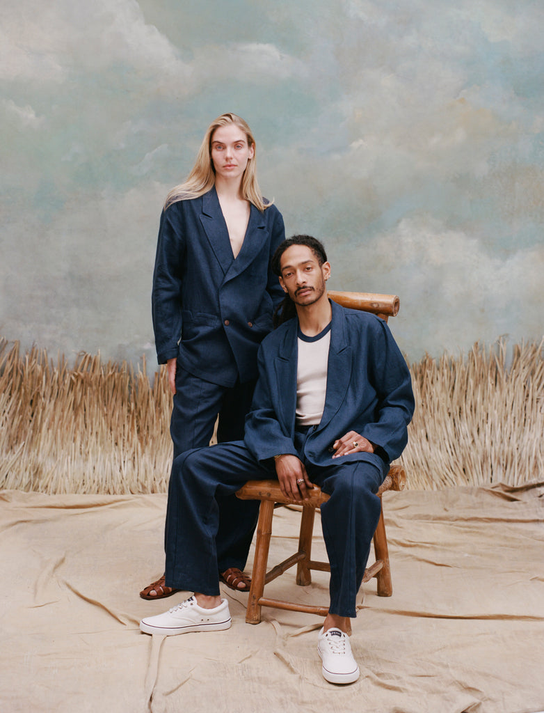 Female model with long-blonde hair next to a seated male model with dreadlocks as they both wear the 'Easy-Going Jacket' in Navy Linen alongside matching navy pants, with the male wearing a white shirt underneath and white sneakers while the female model is wearing nothing underneath the jacket, all shot on a textured light-blue background with waist-high beige grass. 
