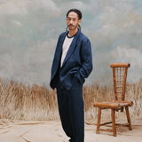 Male model with dreadlocks wearing the 'Easy-Going Jacket' in navy linen with white shirt underneath and matching navy linen pants alongside white shoes, shot on a light blue textured background with waist high beige grass.