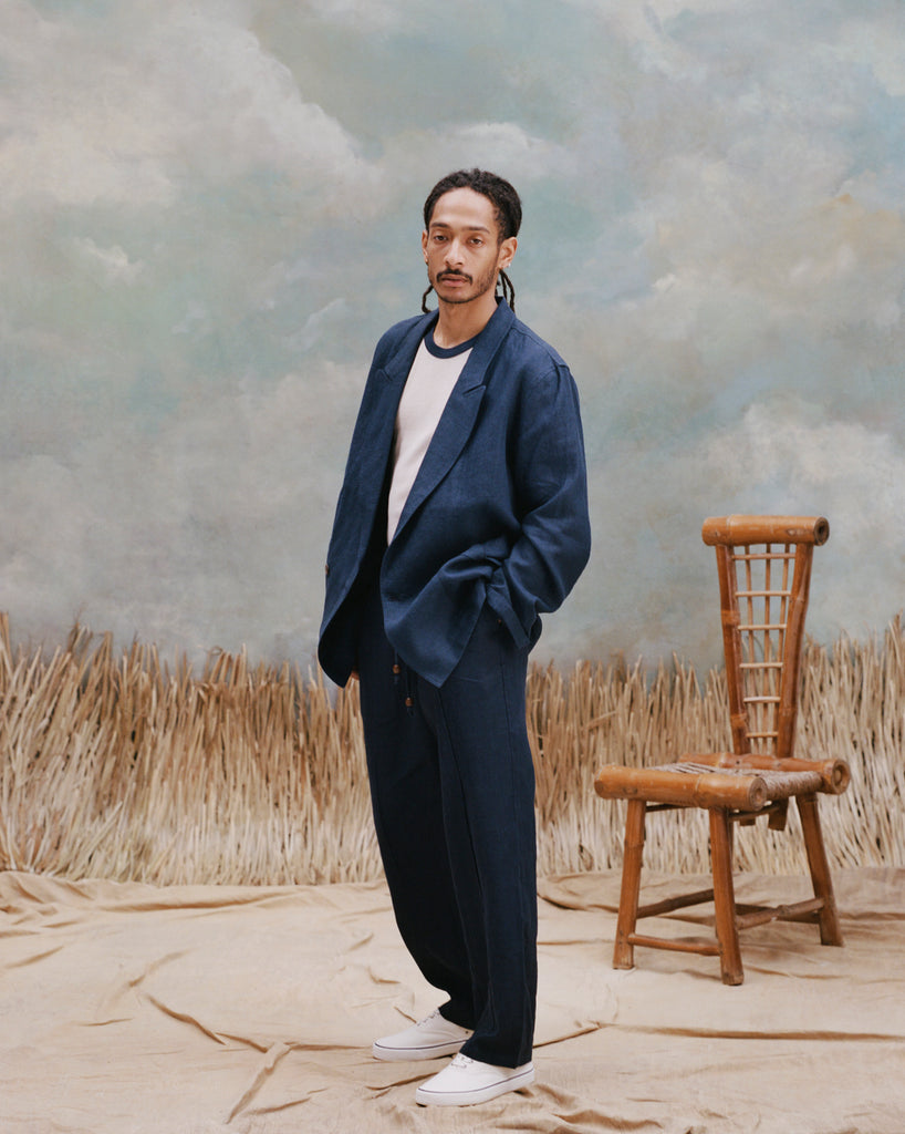 Male model with dreadlocks wearing the 'Easy-Going Jacket' in navy linen with white shirt underneath and matching navy linen pants alongside white shoes, shot on a light blue textured background with waist high beige grass.