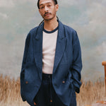 Male model with dreadlocks wearing the 'Easy-Going Jacket' in navy linen with a white shirt that has a navy collar, with matching navy linen pants, shot on a light blue textured background, and beige grass.