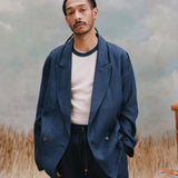 Male model with dreadlocks wearing the 'Easy-Going Jacket' in navy linen with a white shirt that has a navy collar, with matching navy linen pants, shot on a light blue textured background, and beige grass.