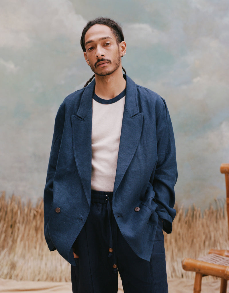 Male model with dreadlocks wearing the 'Easy-Going Jacket' in navy linen with a white shirt that has a navy collar, with matching navy linen pants, shot on a light blue textured background, and beige grass.