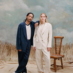 Male model with dreadlocks wearing the 'easy-going jacket' in navy linen unbuttoned with a white shirt that has blue lining on the collar with matching navy linen pants and white vans, resting his hand on the shoulder of a female model wearing the 'easy-going' suit in white with a white shirt underneath and matching white linen pants and white loafesr, shot on a light blue textured background with beige grass.