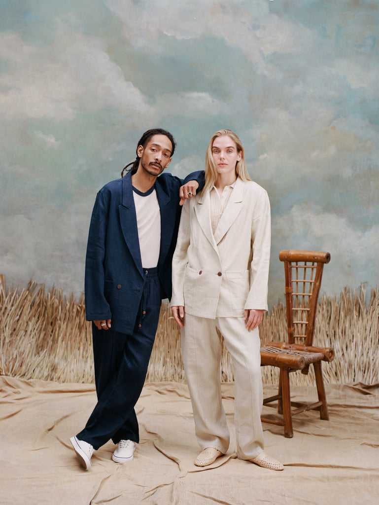 Male model with dreadlocks wearing the 'easy-going jacket' in navy linen unbuttoned with a white shirt that has blue lining on the collar with matching navy linen pants and white vans, resting his hand on the shoulder of a female model wearing the 'easy-going' suit in white with a white shirt underneath and matching white linen pants and white loafesr, shot on a light blue textured background with beige grass.