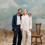 Male model with dreadlocks standing with one arm around the female model with long-blonde hair who is wearing the 'Easy-Going Jacket' in sand linen alongside matching sand linen pants, and beige loafers, while the male model wears a navy suit set with a white shirt underneath, and white shoes, shot against a textured light blue background with waist-high beige grass.