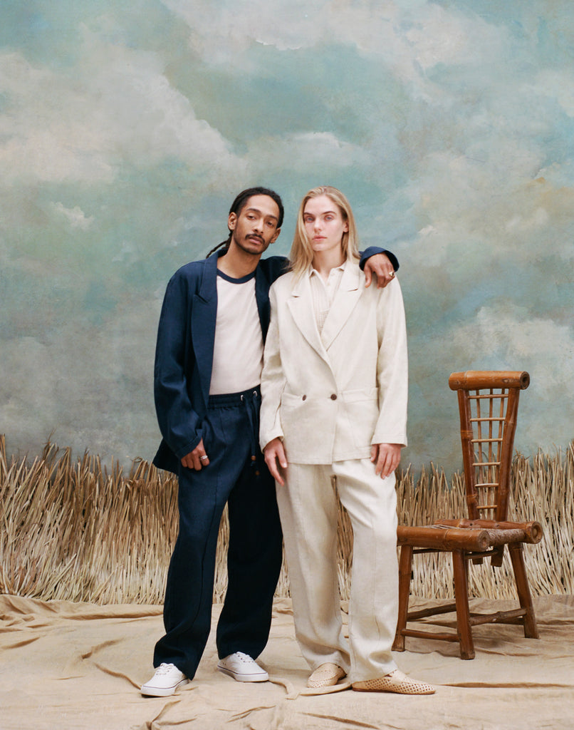 Male model with dreadlocks standing with one arm around the female model with long-blonde hair who is wearing the 'Easy-Going Jacket' in sand linen alongside matching sand linen pants, and beige loafers, while the male model wears a navy suit set with a white shirt underneath, and white shoes, shot against a textured light blue background with waist-high beige grass.