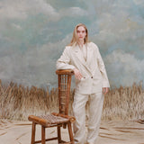Female model with long-blonde hair wearing the 'Easy-Going Jacket' in sand linen with one hand resting on a bamboo chair, with matching sand linen pants, a beige collared shirt, and beige loafers, shot a light blue textured background with waist-high beige grass.