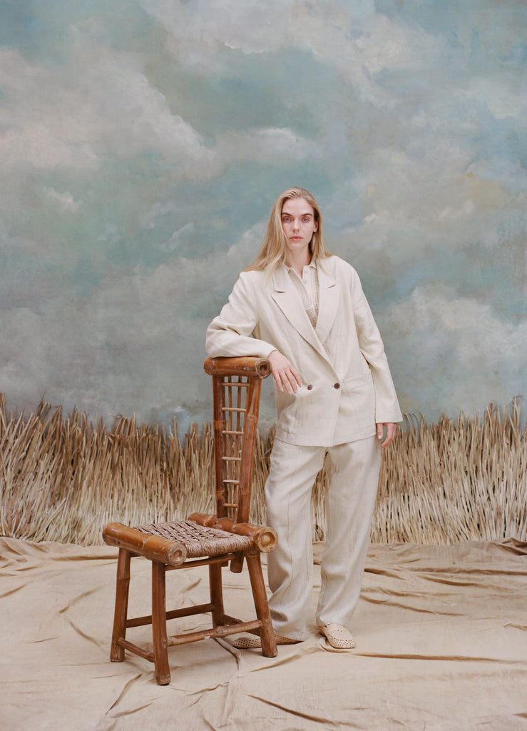 Female model with long-blonde hair wearing the 'Easy-Going Jacket' in sand linen with one hand resting on a bamboo chair, with matching sand linen pants, a beige collared shirt, and beige loafers, shot a light blue textured background with waist-high beige grass.