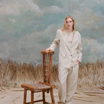 Female model with long blonde hair wearing the 'Easy-Going Pants' in Sand Linen with one hand resting on a bamboo chair and the other in her pocket, while she wears a matching blazer in sand linen with a beige collared shirt, shot on a light blue textured background and waist-high beige grass.