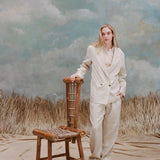 Female model with long blonde hair wearing the 'Easy-Going Pants' in Sand Linen with one hand resting on a bamboo chair and the other in her pocket, while she wears a matching blazer in sand linen with a beige collared shirt, shot on a light blue textured background and waist-high beige grass.