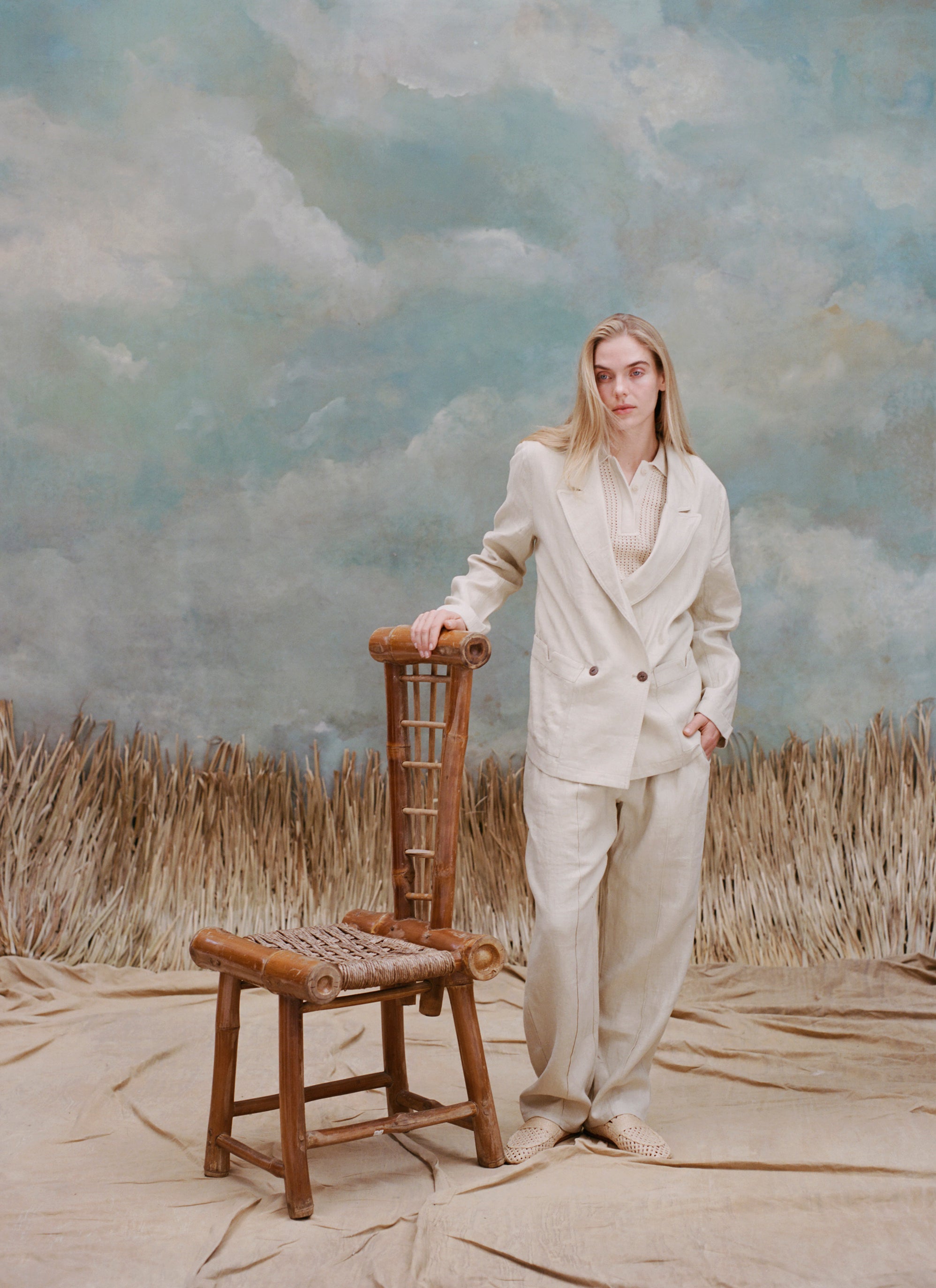 Female model with long blonde hair wearing the 'Easy-Going Pants' in Sand Linen with one hand resting on a bamboo chair and the other in her pocket, while she wears a matching blazer in sand linen with a beige collared shirt, shot on a light blue textured background and waist-high beige grass.