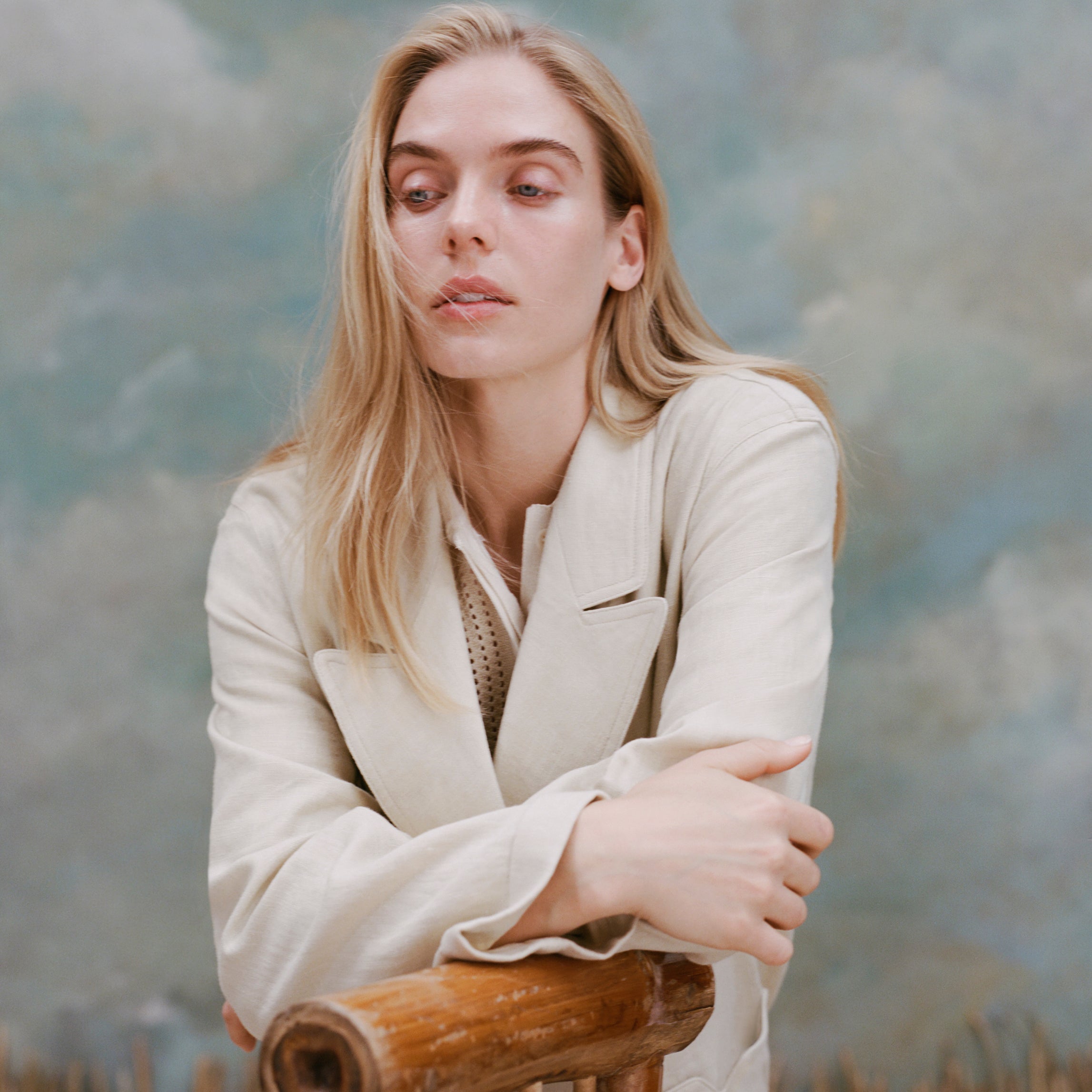 Female model with long-blonde hair wearing the 'Easy-Going Jacket' in sand linen with her hands crossed resting on a bamboo chair, shot on a textured light blue background.