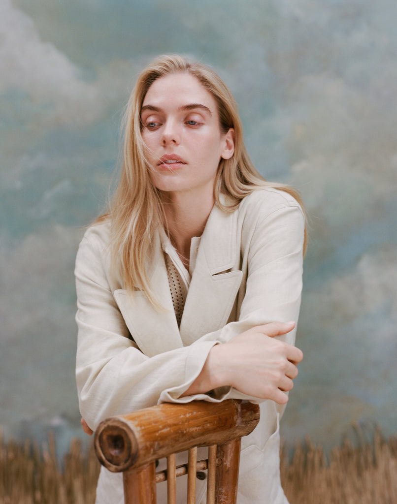 Female model with long-blonde hair wearing the 'Easy-Going Jacket' in sand linen with her hands crossed resting on a bamboo chair, shot on a textured light blue background.